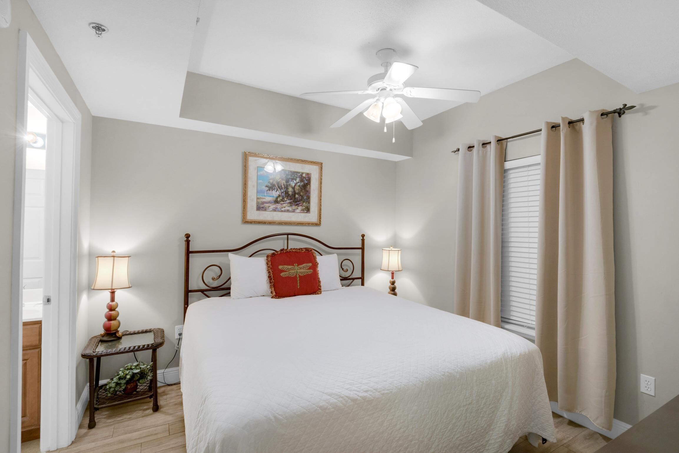 A bedroom with a bed, two bedside lamps, a ceiling fan, a framed picture on the wall, and a window with curtains.
