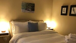 Ein ordentlich bezogenes Bett mit weißen Leinen und einem blauen Kissen, zwei Nachttischlampen und ein gerahmtes Bild an der Wand.
