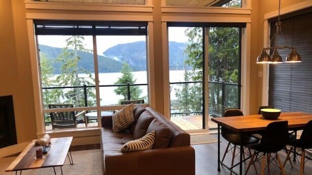 Ein gemütliches Wohnzimmer mit einer Couch, einem Esstisch und Stühlen. Große Fenster bieten Blick auf einen See und Berge.