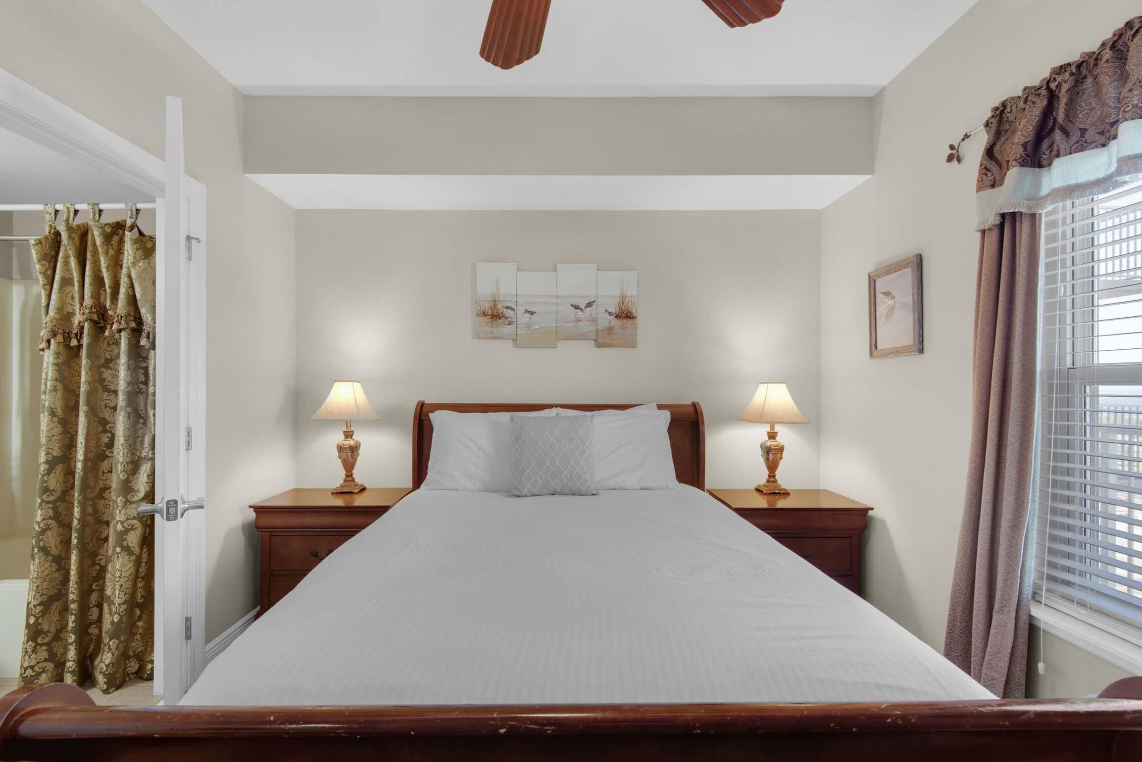 A neatly made bed with a headboard, two bedside lamps, a wall-mounted artwork, and a window with curtains.