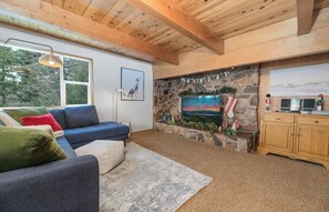 A cozy living room with a stone fireplace, a large window, and a wooden ceiling. - Big Bear Treehouse-HOT TUB-Gorgeous Views (Big Bear Lake)