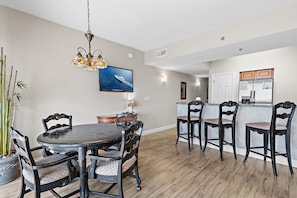 A dining area with a round table and chairs, a television mounted on the wall, and a kitchen area with cabinets and a refrigerator. - Ariel Dunes II 1805 (Miramar Beach)
