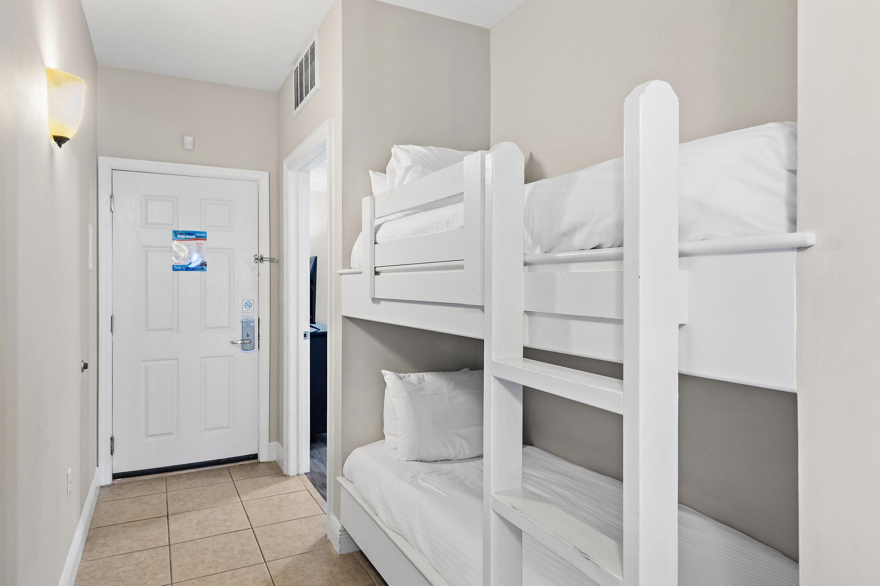 A hotel room with bunk beds, a door with a lock, and a light fixture on the wall.