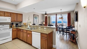 Cozinha moderna com bancadas de granito, armários de madeira e lava-louças. Há uma área de jantar com mesa e cadeiras, e uma grande janela com vista para a cidade.