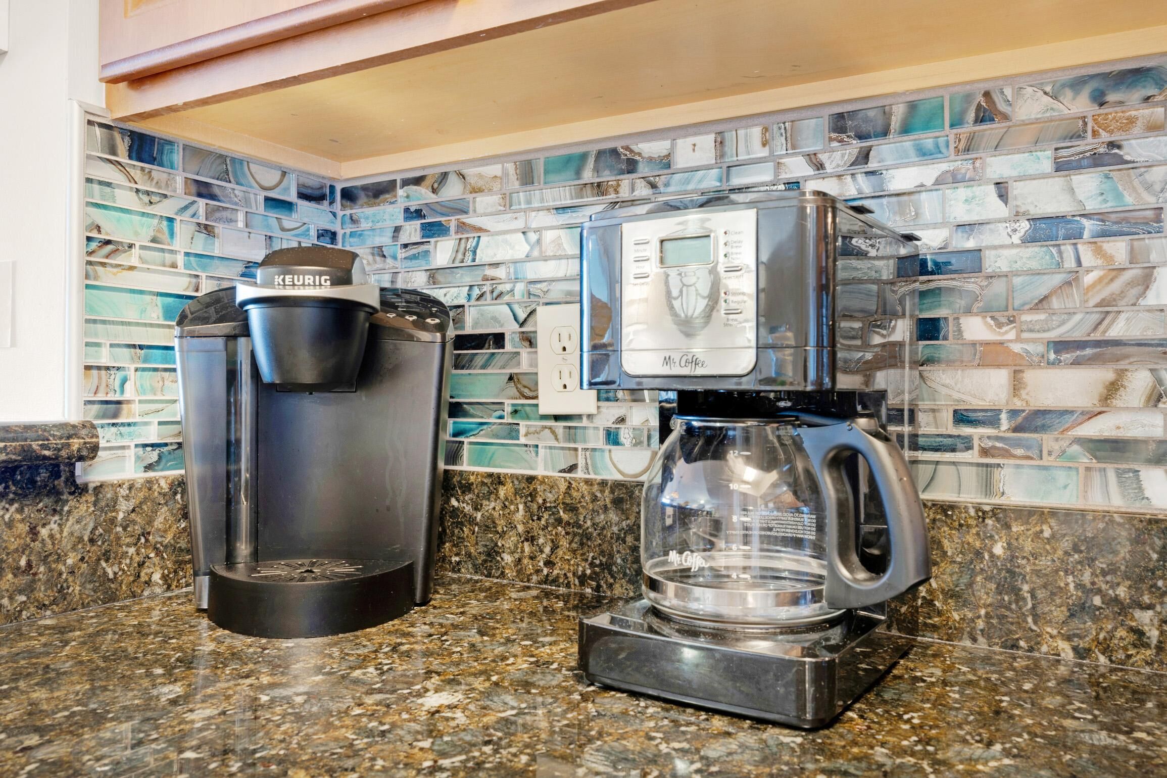 A coffee maker and a glass carafe on a granite countertop with a mosaic backsplash.