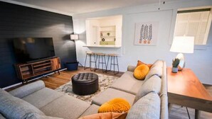 A modern living room with a sofa, ottoman, and a TV. There is a kitchen area with stools and a counter. - Spacious, Town Square, Theatre Room! 4 bed / 2 bath Private Home on town square (Covington)