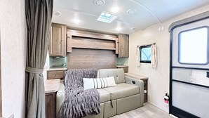 A cozy RV interior with a sofa bed, wooden cabinets, and a small window. - Joy ~ The Happy Little Camper (Loranger)