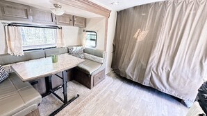 A compact RV interior with a kitchenette, seating area, and a curtained window. - Joy ~ The Happy Little Camper (Loranger)
