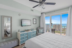 A bedroom with a large bed, a ceiling fan, a TV, a dresser, and a balcony with a view of the city.