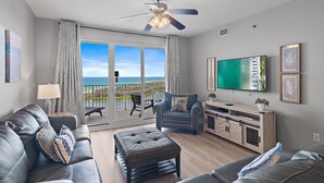 Un salon moderne avec un canapé en cuir, un pouf capitonné, un téléviseur à écran plat et un balcon offrant une vue sur l’océan.