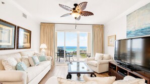 A spacious living room with a large TV, a ceiling fan, and a balcony with a view of the ocean. - Ariel Dunes II 1509 (Miramar Beach)