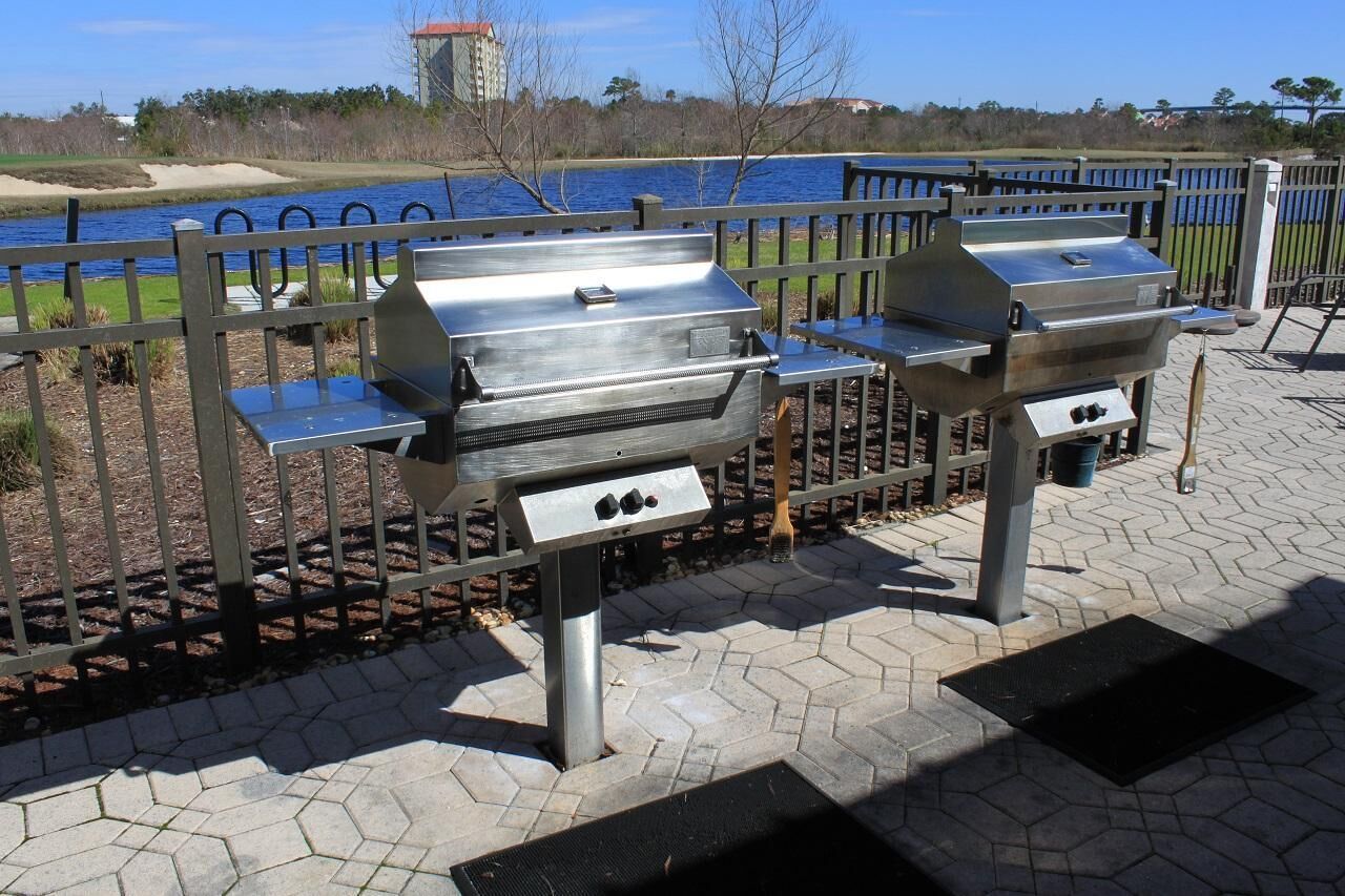 Two portable gas grills on a paved patio with a view of a body of water and a wooden fence.