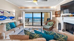 A spacious living room with a large sofa, a coffee table, and a fireplace. There is a television mounted on the wall and a ceiling fan. The room has a large window with a view of the ocean. - Shipwatch D604 (Pensacola)