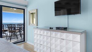 A room with a large TV mounted on the wall, a glass-top dresser, and a view of the ocean through a sliding glass door. - Shipwatch D604 (Pensacola)