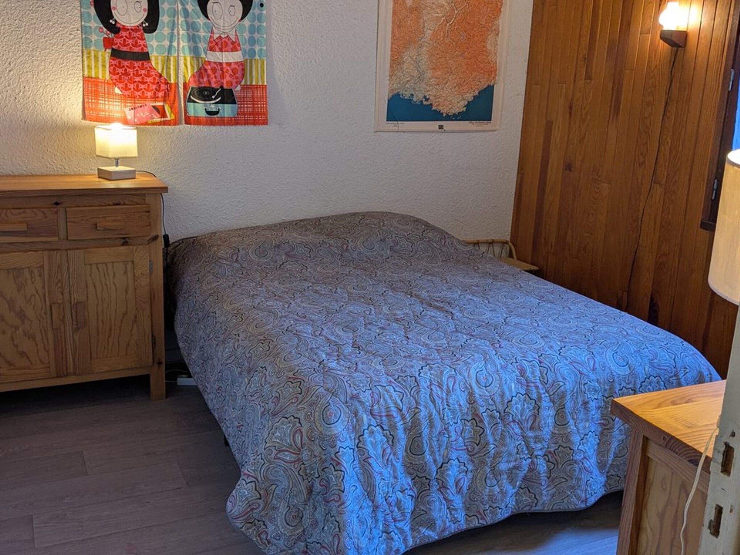 A bedroom with a patterned bedspread, wooden furniture, and wall decorations including a map and cartoon characters.