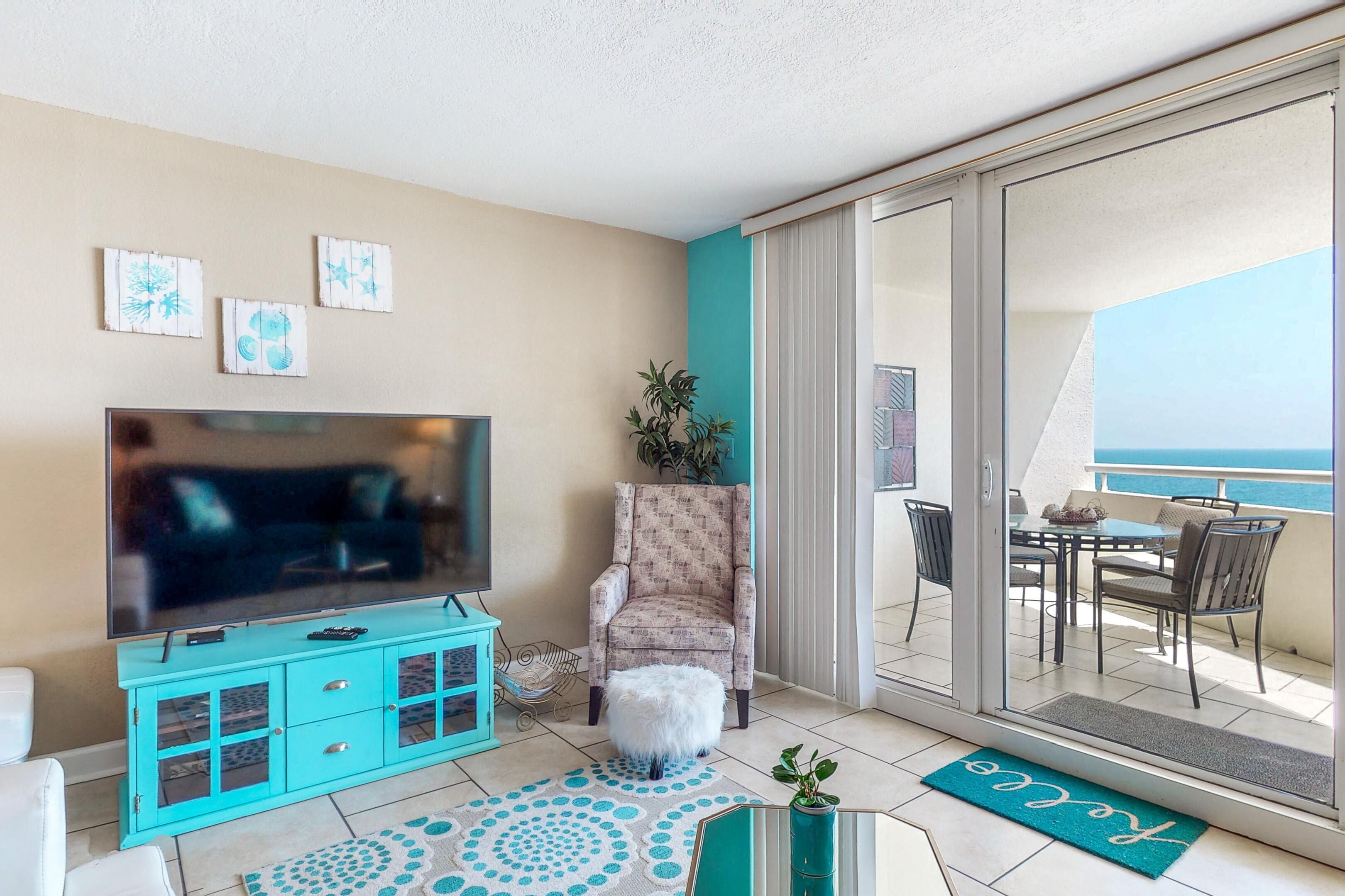 Een woonkamer met een flatscreen-tv, een gestreept fauteuil, een klein wit krukje en uitzicht op een balkon met buitenmeubilair.