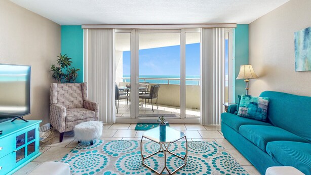 A living room with a TV, a teal sofa, a patterned armchair, and a view of the ocean through a sliding door. - Perdido Sun 612 (Pensacola)
