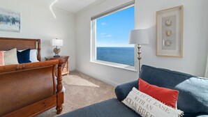 A bedroom with a large window, a bed, a nightstand, a lamp, and a framed picture on the wall. - Indigo West 1802 (Pensacola)
