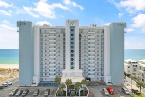 A large hotel with multiple floors, a central tower, and a parking lot with several cars. - Summerchase 905 (Orange Beach)