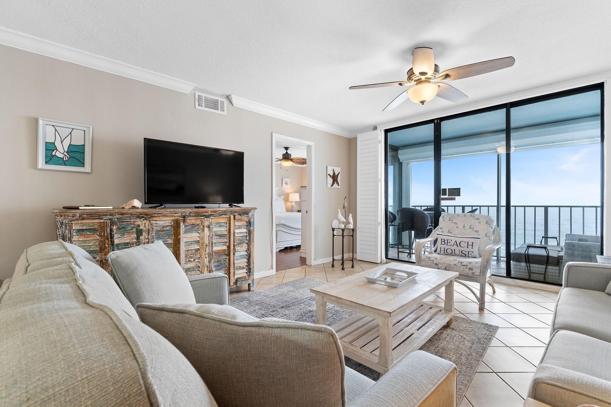 A spacious living room with a large flat-screen TV, comfortable seating, a ceiling fan, and a view of the ocean through large glass doors.