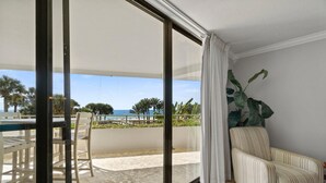 A room with a large sliding glass door leading to a balcony with a view of the sea and palm trees. There is a striped armchair and a table with chairs on the balcony. - Eden East 101 (Pensacola)