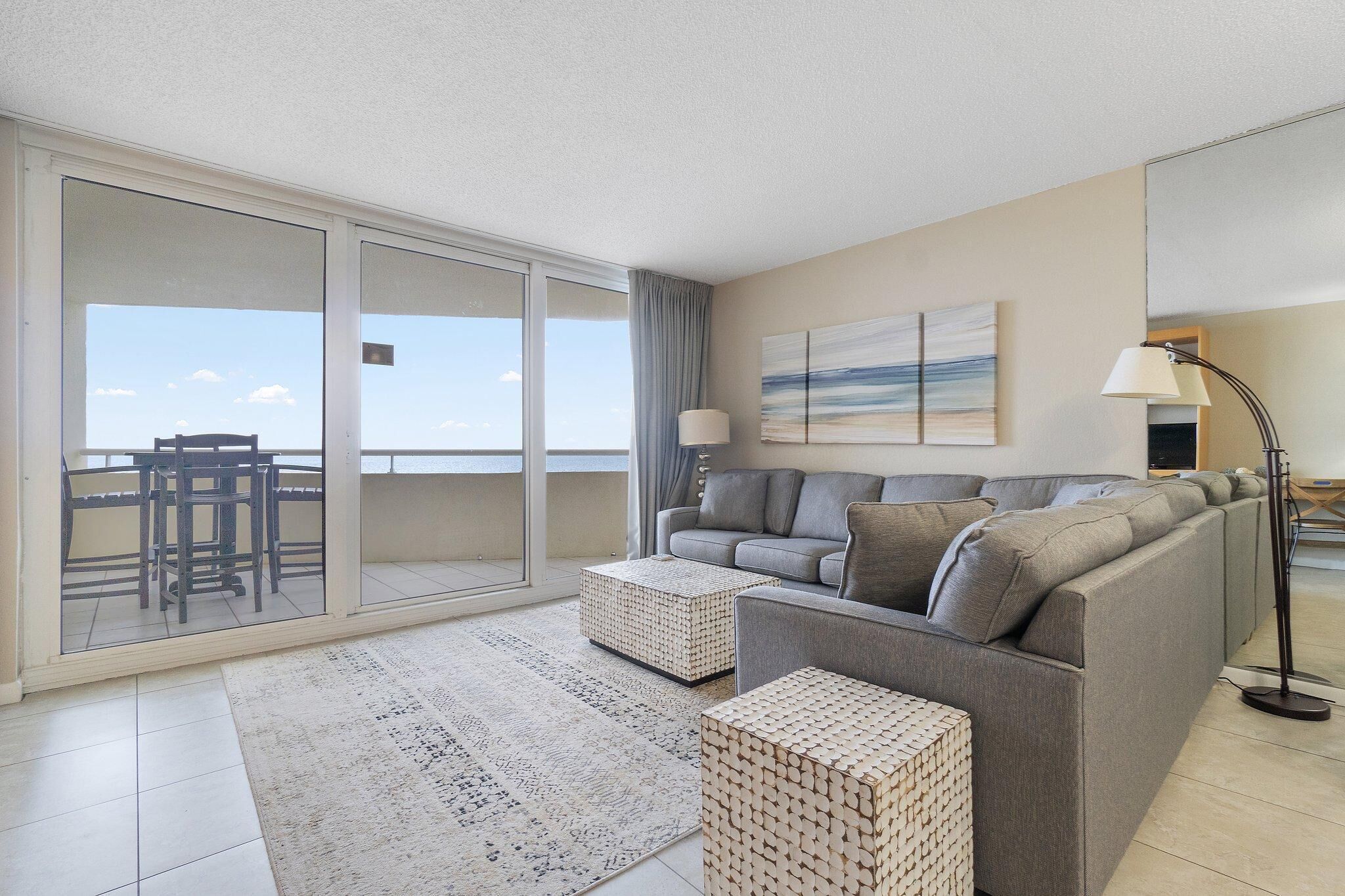 A modern living room with a grey sofa, two woven ottomans, a patterned rug, a floor lamp, and a large sliding glass door leading to a balcony with a view.