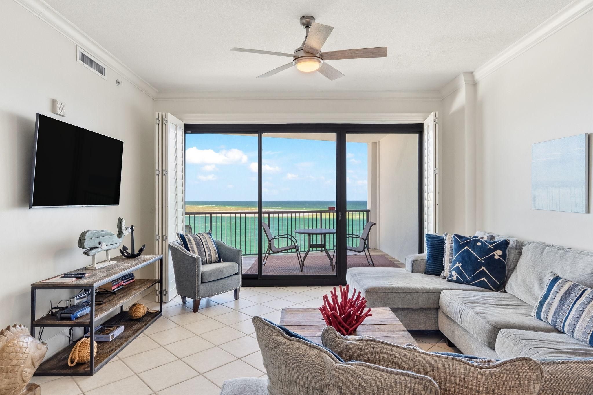 A living room with a large flat-screen TV, a ceiling fan, and a view of the ocean through a sliding glass door.