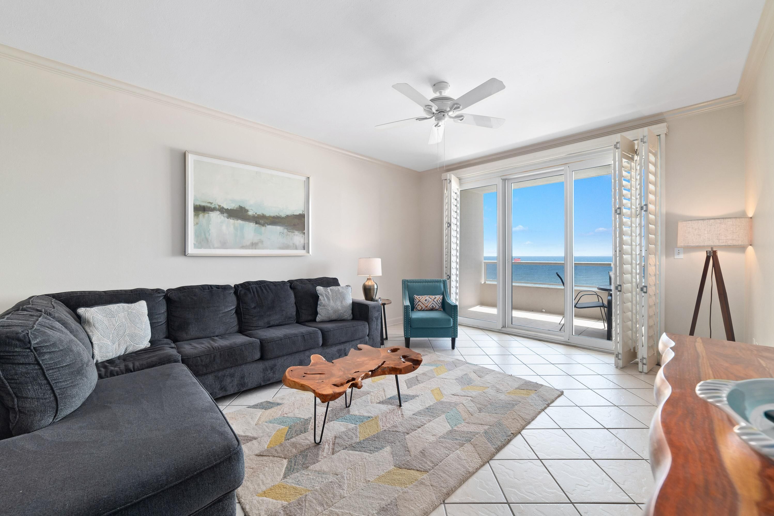 A living room with a dark gray sectional sofa, a wooden coffee table, a teal armchair, and a view of the ocean through large glass doors.