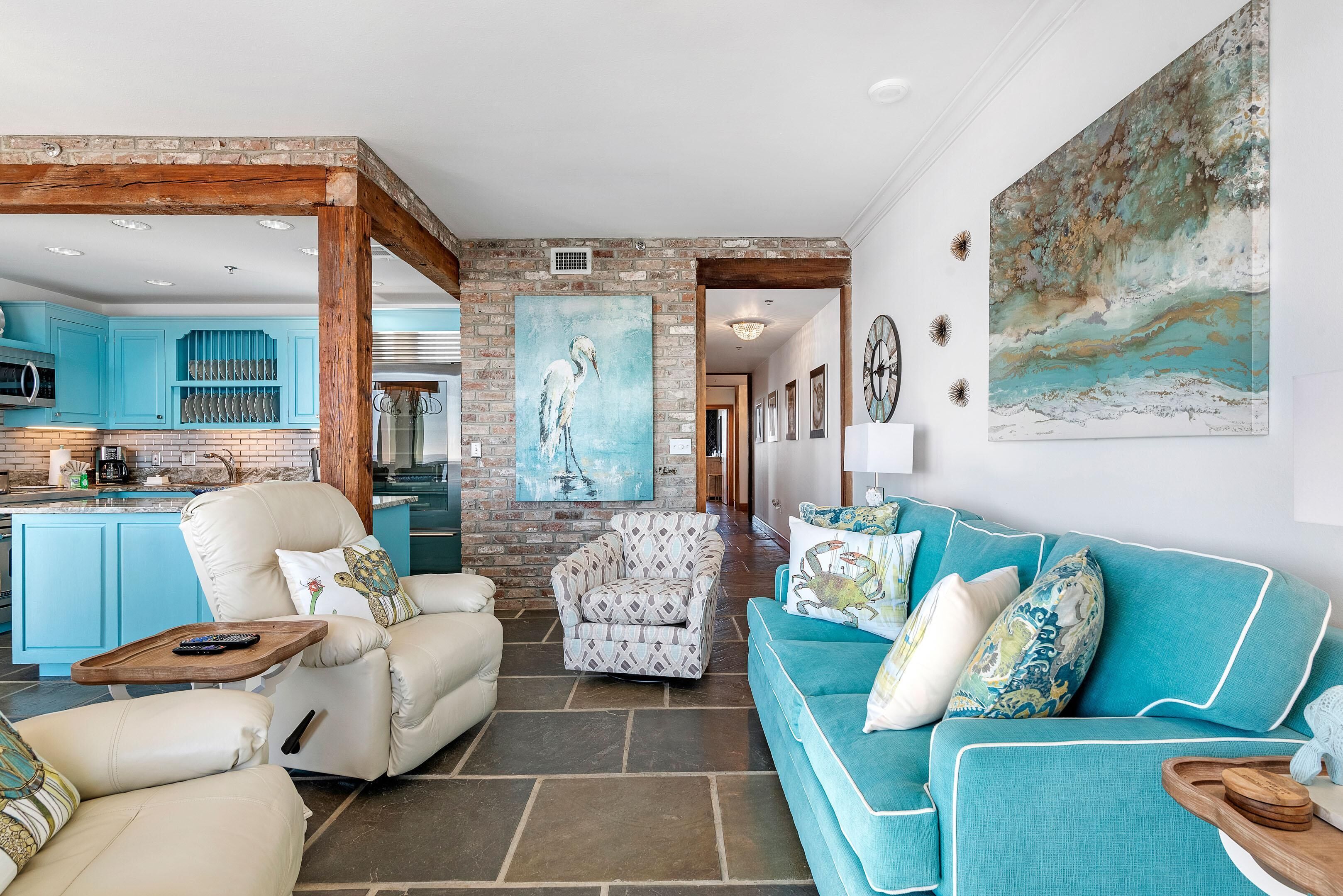 A spacious living area with a kitchen in the background, featuring a teal sofa, a white armchair, and a patterned armchair.