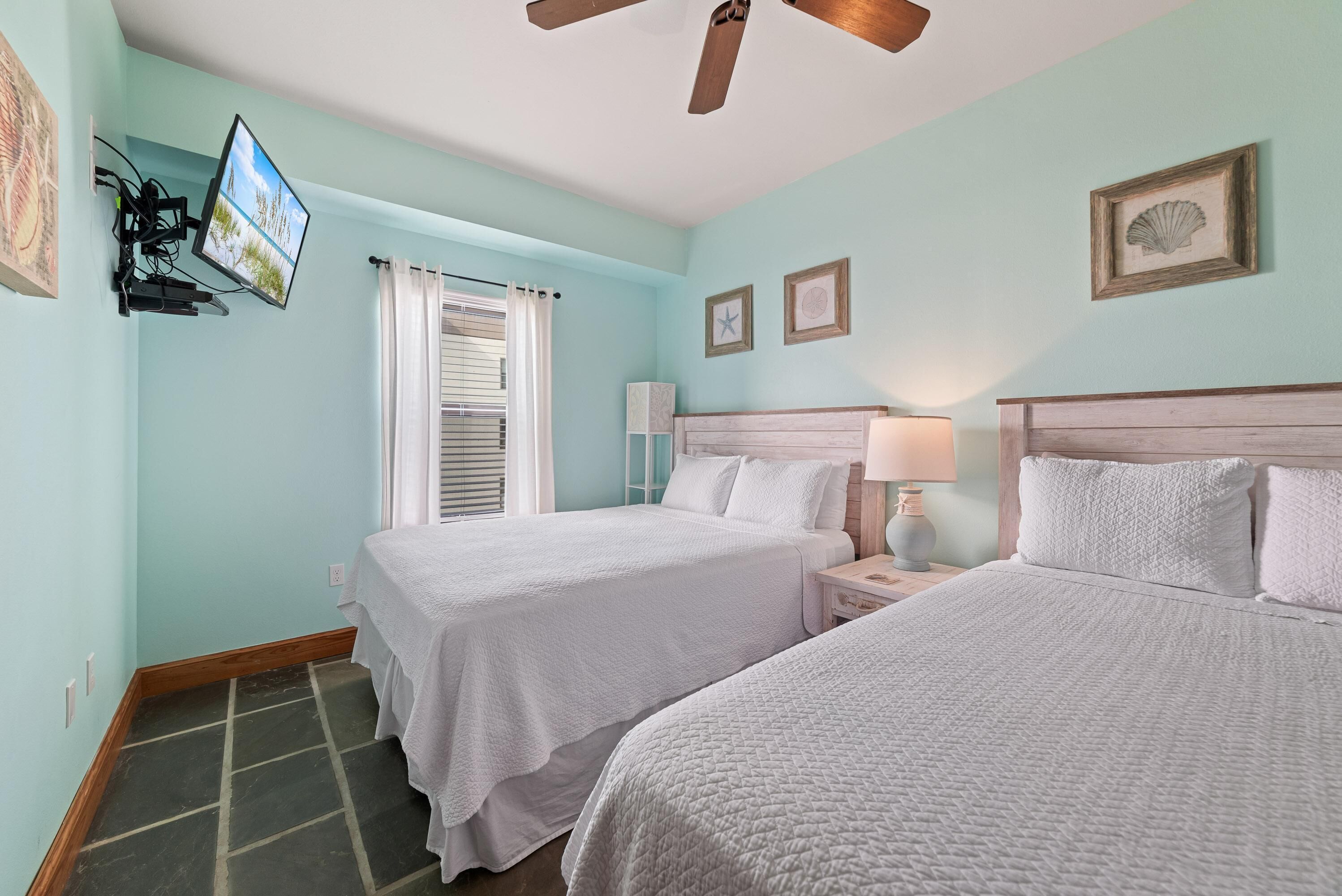 A room with two beds, a ceiling fan, a TV mounted on the wall, and a window with curtains.