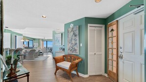 A spacious living area with a wooden bench, a glass coffee table, and a dining table set with chairs. There is a large painting on the wall and a wooden door with a glass panel.