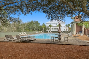 A swimming pool with lounge chairs and a deck area, surrounded by trees and a large house in the background. - Starfish Cottage (Orange Beach)