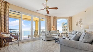 A spacious living room with a large sofa, a smaller sofa, a coffee table, and a wicker chair. There is a ceiling fan, a large window with curtains, and a view of the outside. - Beach Colony Tower Penthouse 17C (Pensacola)
