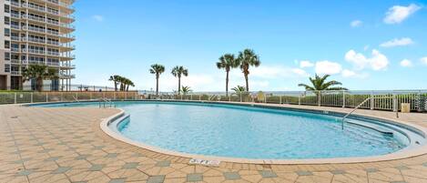A swimming pool with a tiled deck, surrounded by palm trees and a high-rise building.