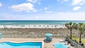 A pool area with lounge chairs and umbrellas, overlooking a beach with umbrellas and people. - Sandy Key 713 (Pensacola)