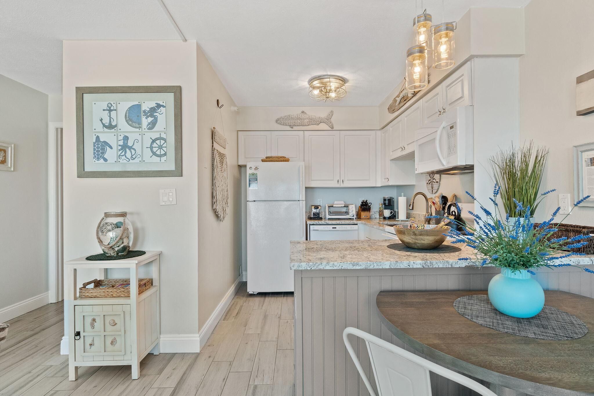 A kitchen with white cabinets, a refrigerator, a dining area with a table and chairs, and a wall with nautical-themed artwork.