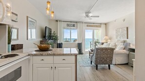 A modern kitchen with white cabinets, a granite countertop, and a dining area with a patterned chair and a white couch. - Perdido Skye 65 (Pensacola)