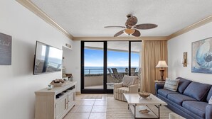 A living room with a view of the ocean, a ceiling fan, a sofa, a coffee table, and a TV on a stand. - Eden West 504 (Pensacola)