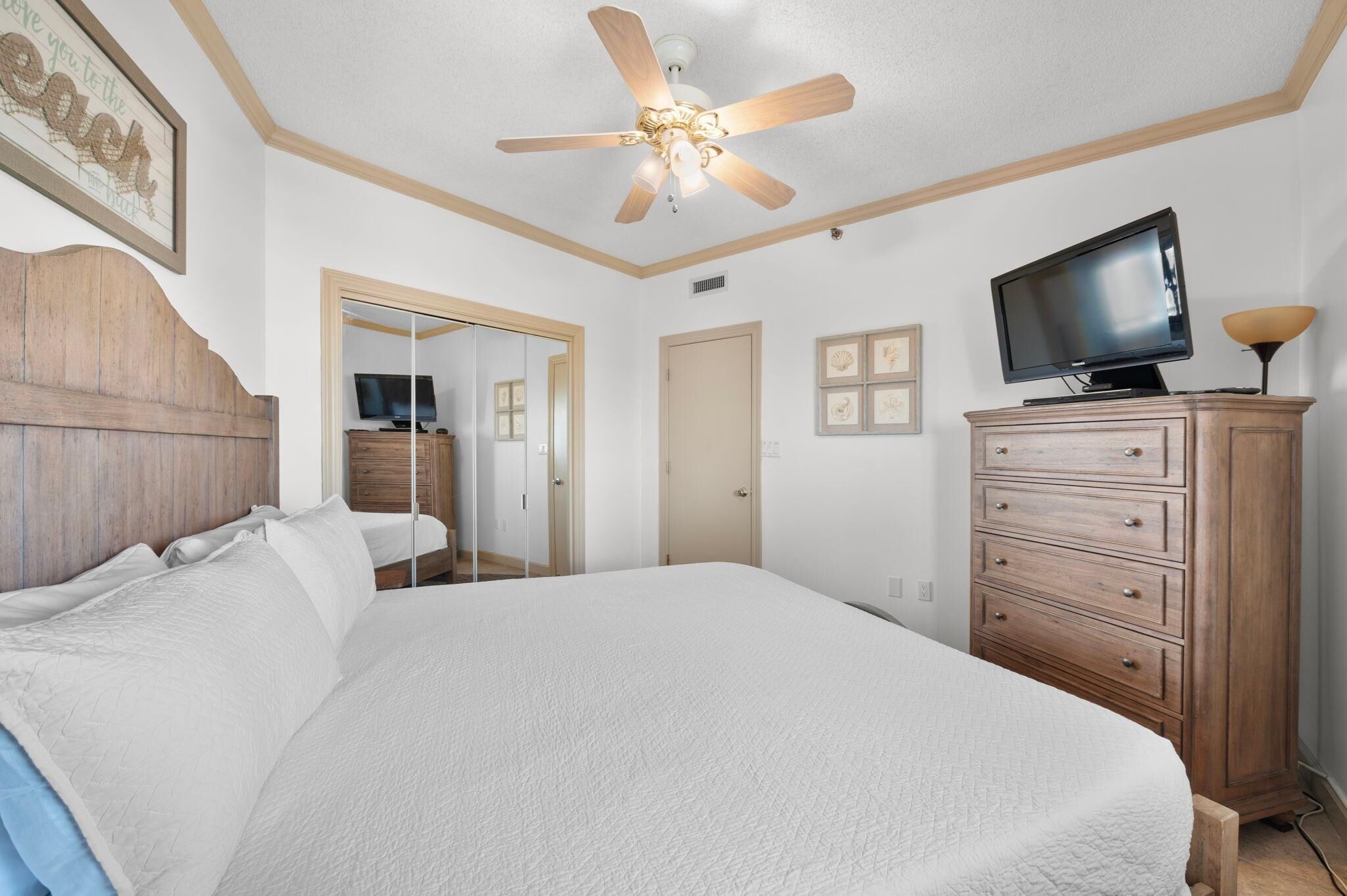 A bedroom with a large bed, a wooden headboard, a ceiling fan, a TV on a wooden cabinet, and a mirror in the background.