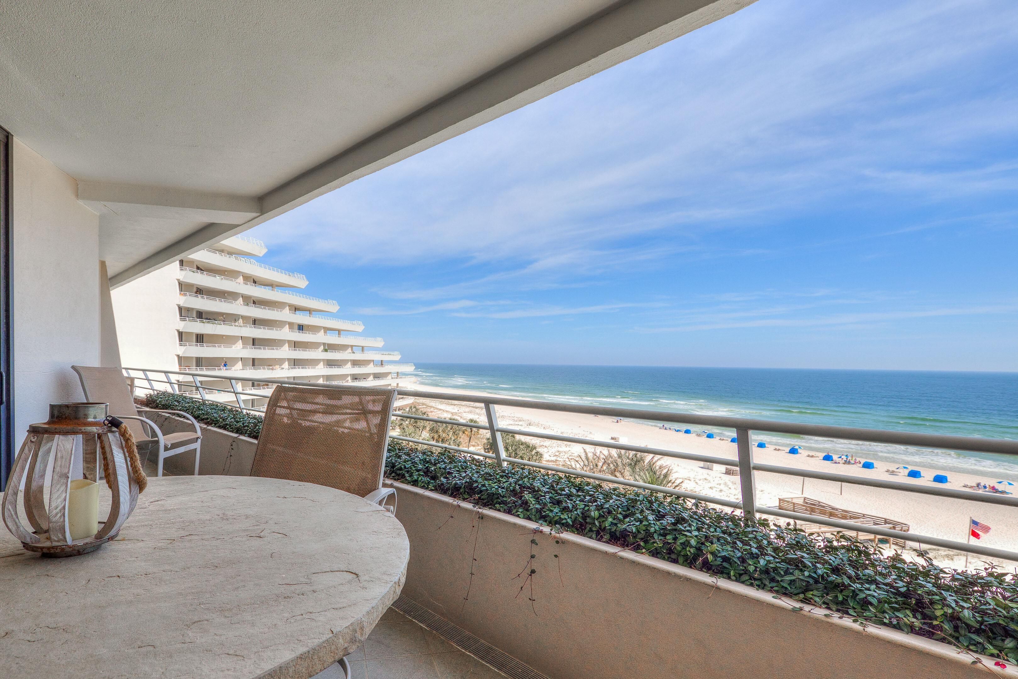 A balcony with a table and chairs, overlooking a beach with umbrellas and a view of the ocean.