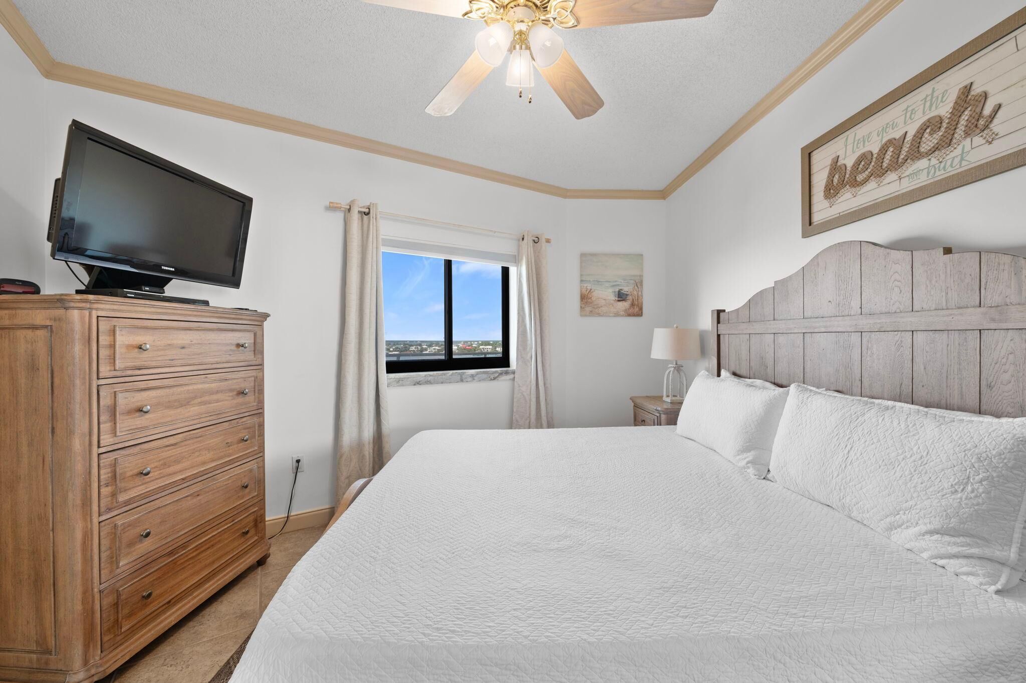 A bedroom with a bed, a television on a wooden dresser, a window with curtains, a ceiling fan, and a wall-mounted sign.