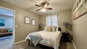 A bedroom with a bed, a ceiling fan, two framed pictures, a nightstand, a lamp, and a painting on the wall. - Near Wichita Airport! Pet Friendly W/ Fenced Patio (Wichita)