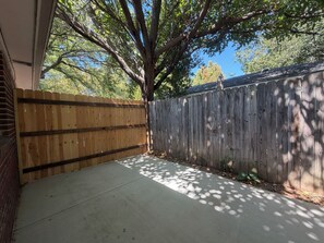A backyard with a wooden fence, a concrete patio, and a tree providing shade.