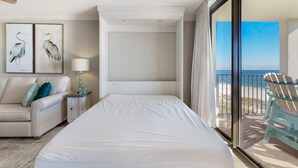 A hotel room with a bed, a nightstand, a lamp, a sofa, and a view of the beach through a sliding door. - Phoenix VII 1002 (Orange Beach)