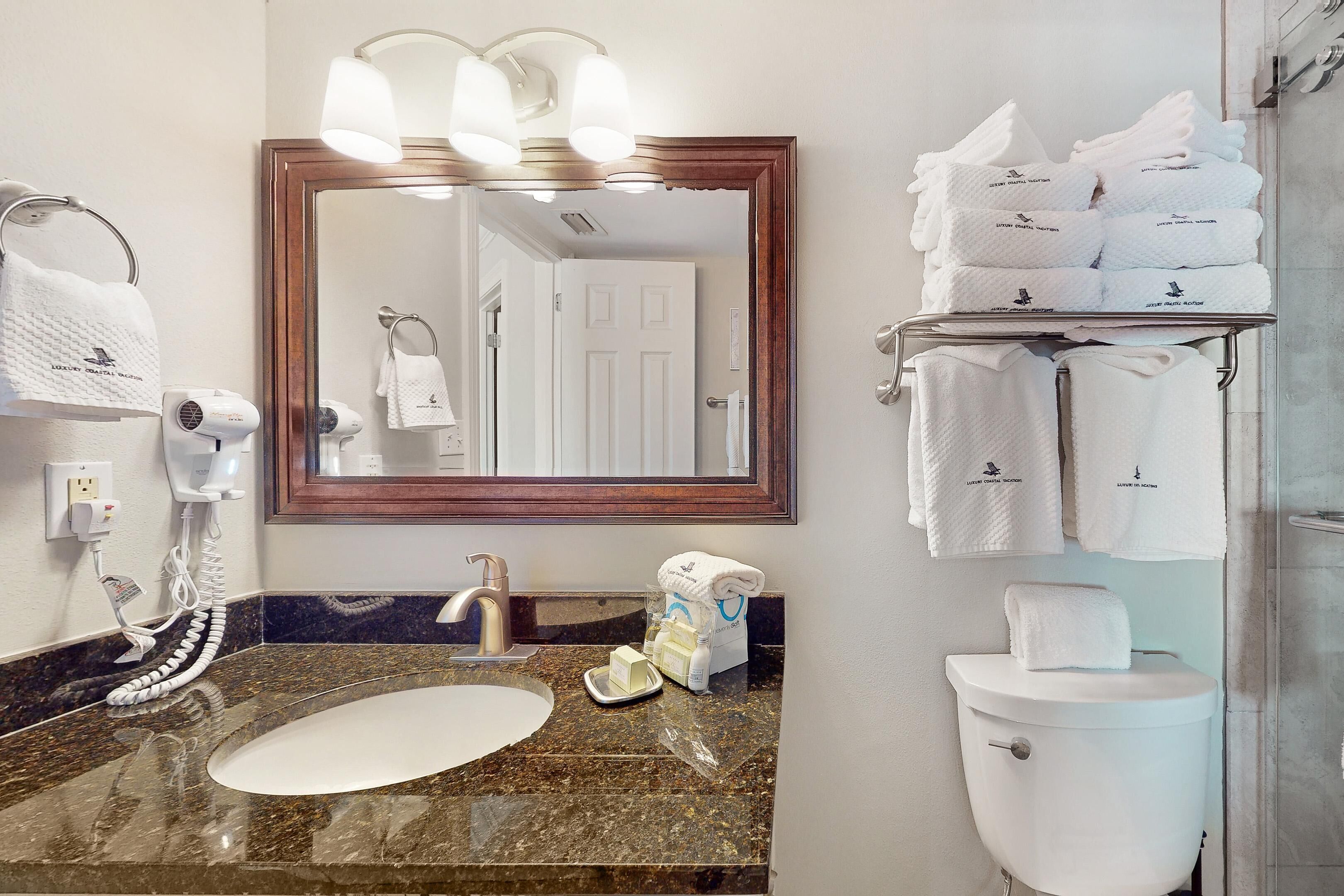 Une salle de bain d’hôtel avec un lavabo en marbre, un grand miroir, un porte-serviettes avec plusieurs serviettes, des toilettes et un sèche-cheveux.