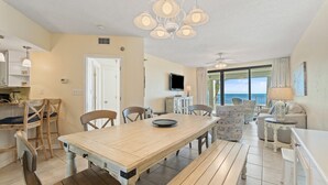 A spacious dining area with a wooden table and chairs, a ceiling fan, and a view of the ocean through large windows. - Pelican Pointe 202 (Orange Beach)