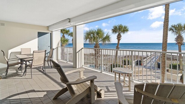 A balcony with a table and chairs, overlooking a beach with palm trees and a clear sky. - Pelican Pointe 202 (Orange Beach)
