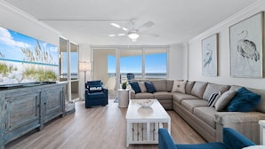 A spacious living room with a large window, a ceiling fan, a blue armchair, a beige sofa, and a white coffee table. - Perdido Sun 416 (Pensacola)