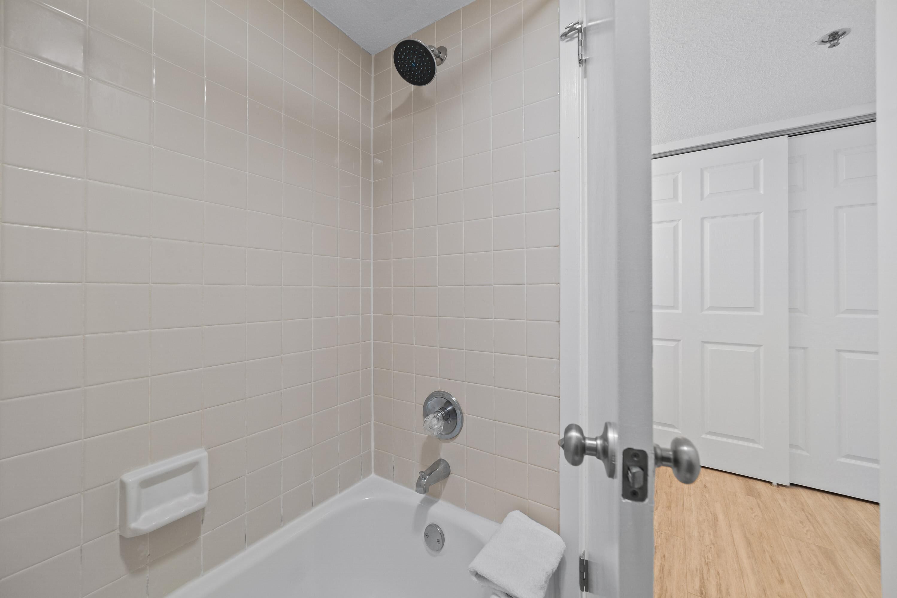 A bathroom with white tiled walls, a bathtub, a showerhead, and a door leading to a hallway.