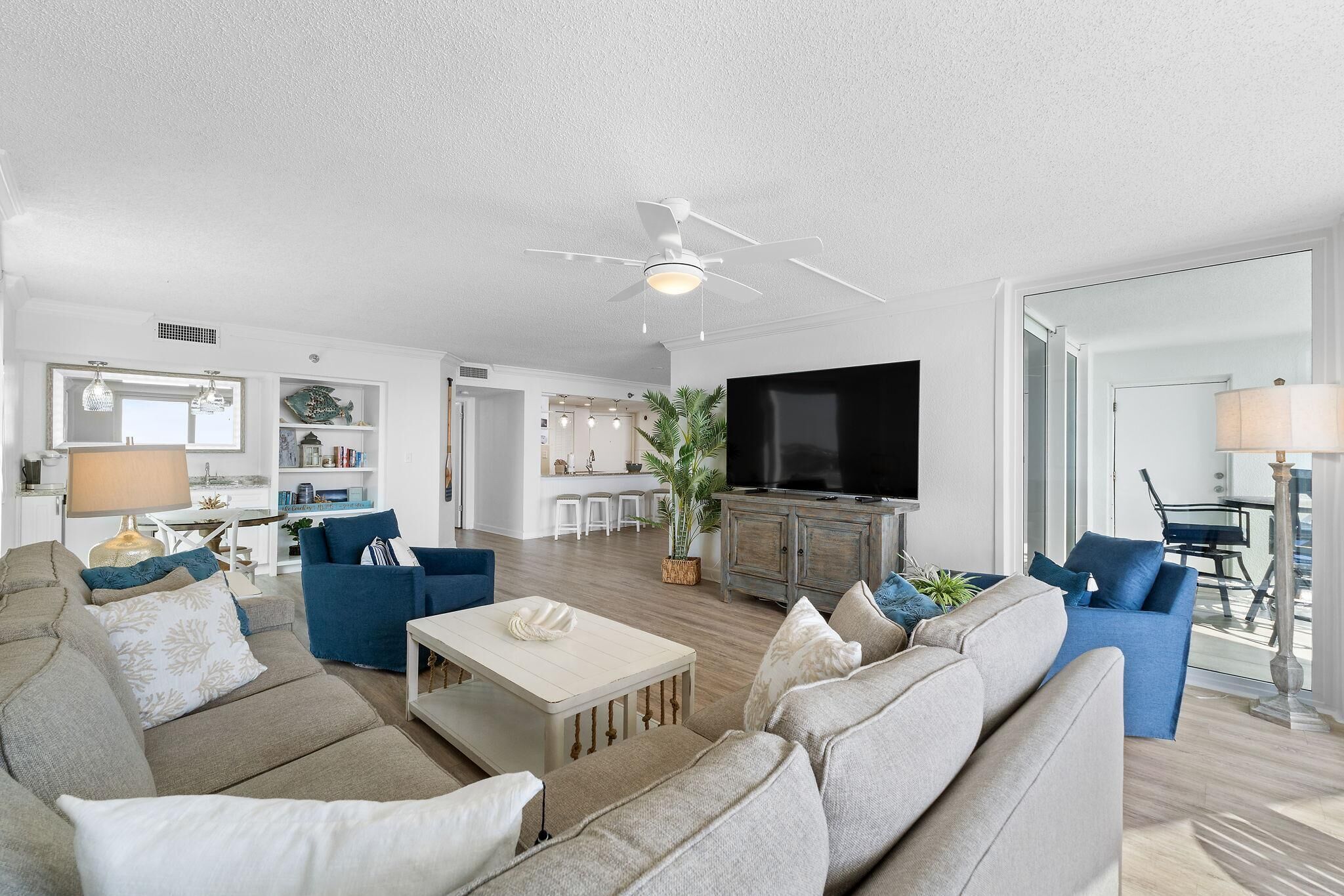 A spacious living room with a ceiling fan, a flat-screen TV, and a kitchen area in the background.